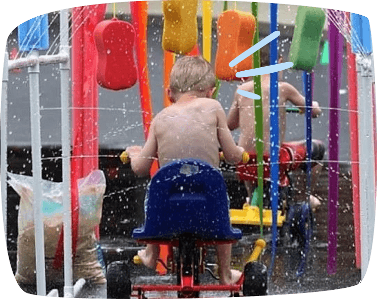 A young shirtless child rides a tricycle through a colorful water splash pad, surrounded by spraying water and bright hanging toys.