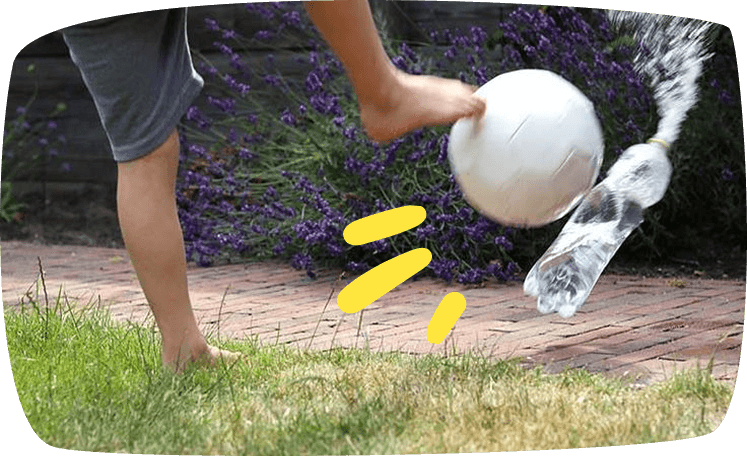 A barefoot person kicks a white ball outdoors on a brick path, sending a plastic bottle flying, with purple lavender bushes in the background.